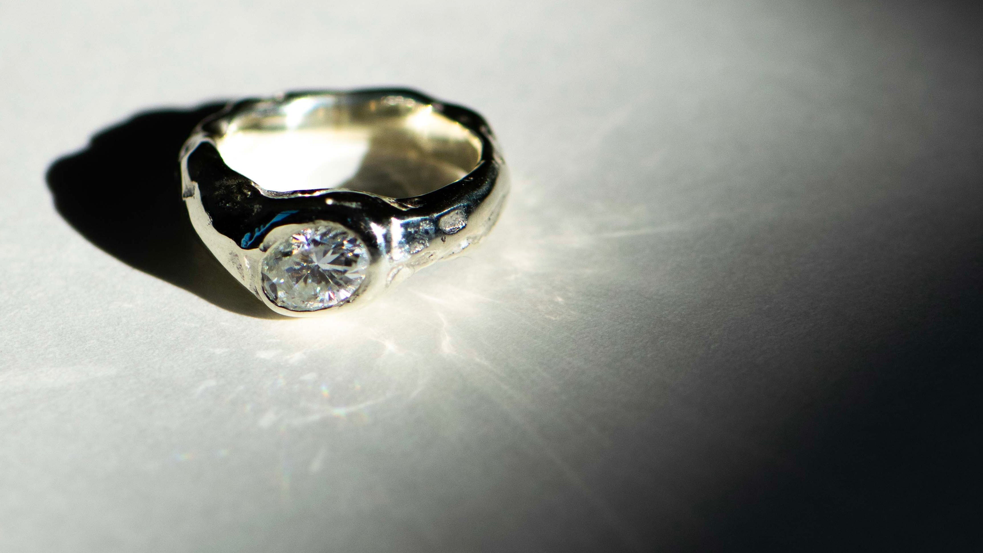 A silver ring with a round clear gemstone, photographed on a white surface under dramatic direct lighting with a hard shadow.