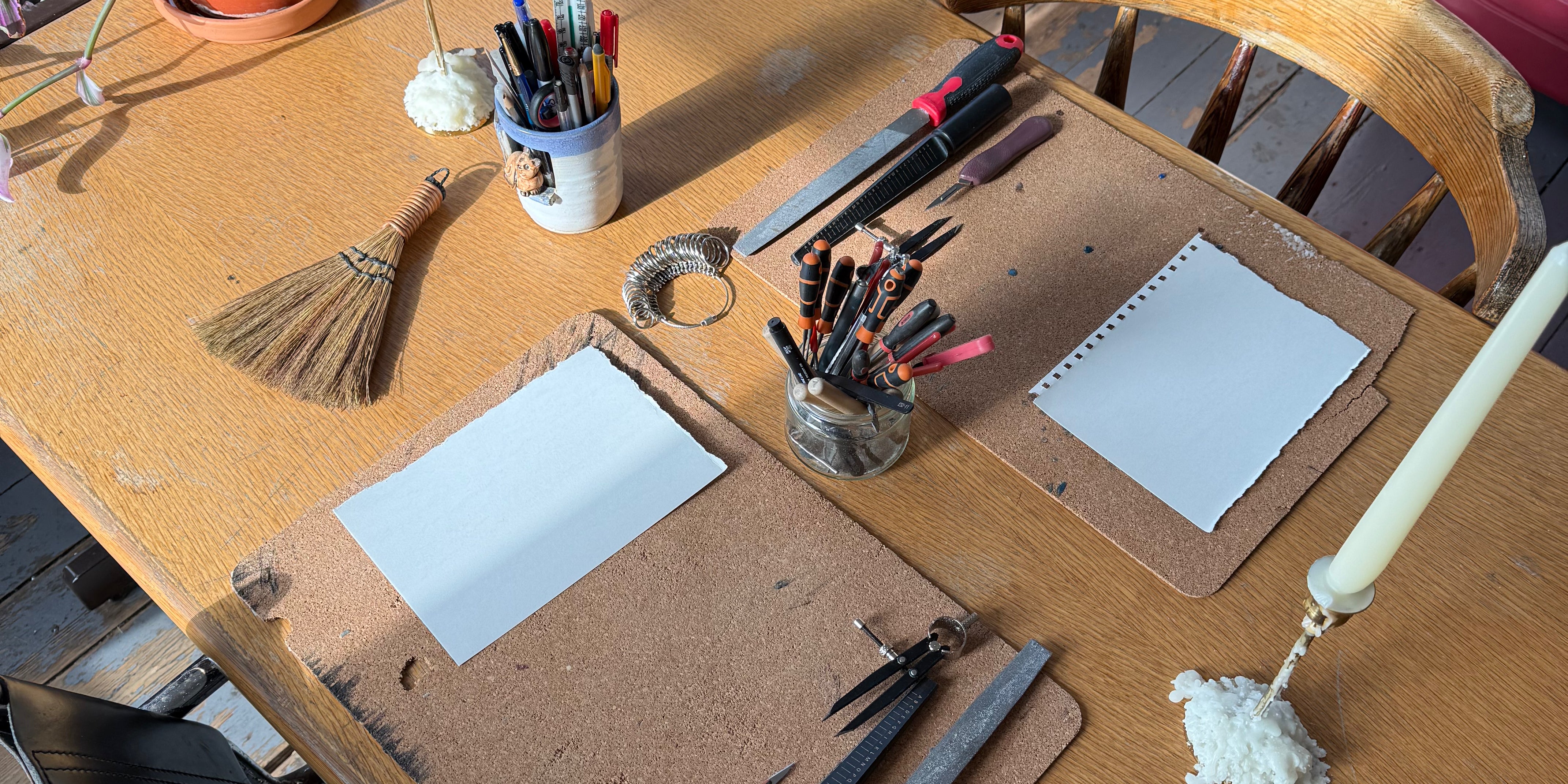 A wooden table in a real room setting, styled with drawing and crafting tools including cardboard cardstock pads, white paper sheets, blue highlighters, a small broom, pencils, markers, and candles. Office chairs, a desk, and other room elements are visible in the background.