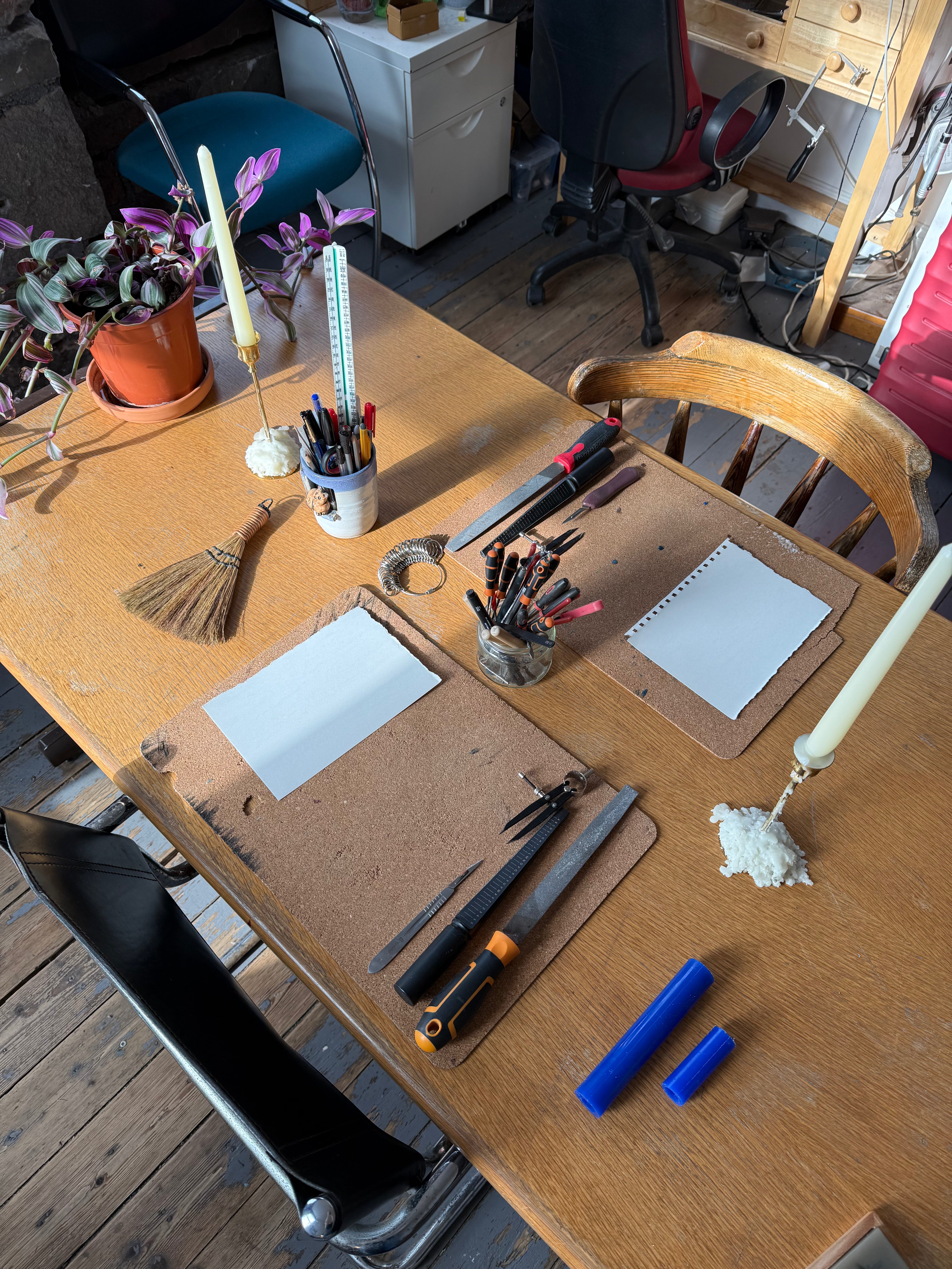 A wooden table in a real room setting, styled with drawing and crafting tools including cardboard cardstock pads, white paper sheets, blue highlighters, a small broom, pencils, markers, and candles. Office chairs, a desk, and other room elements are visible in the background.
