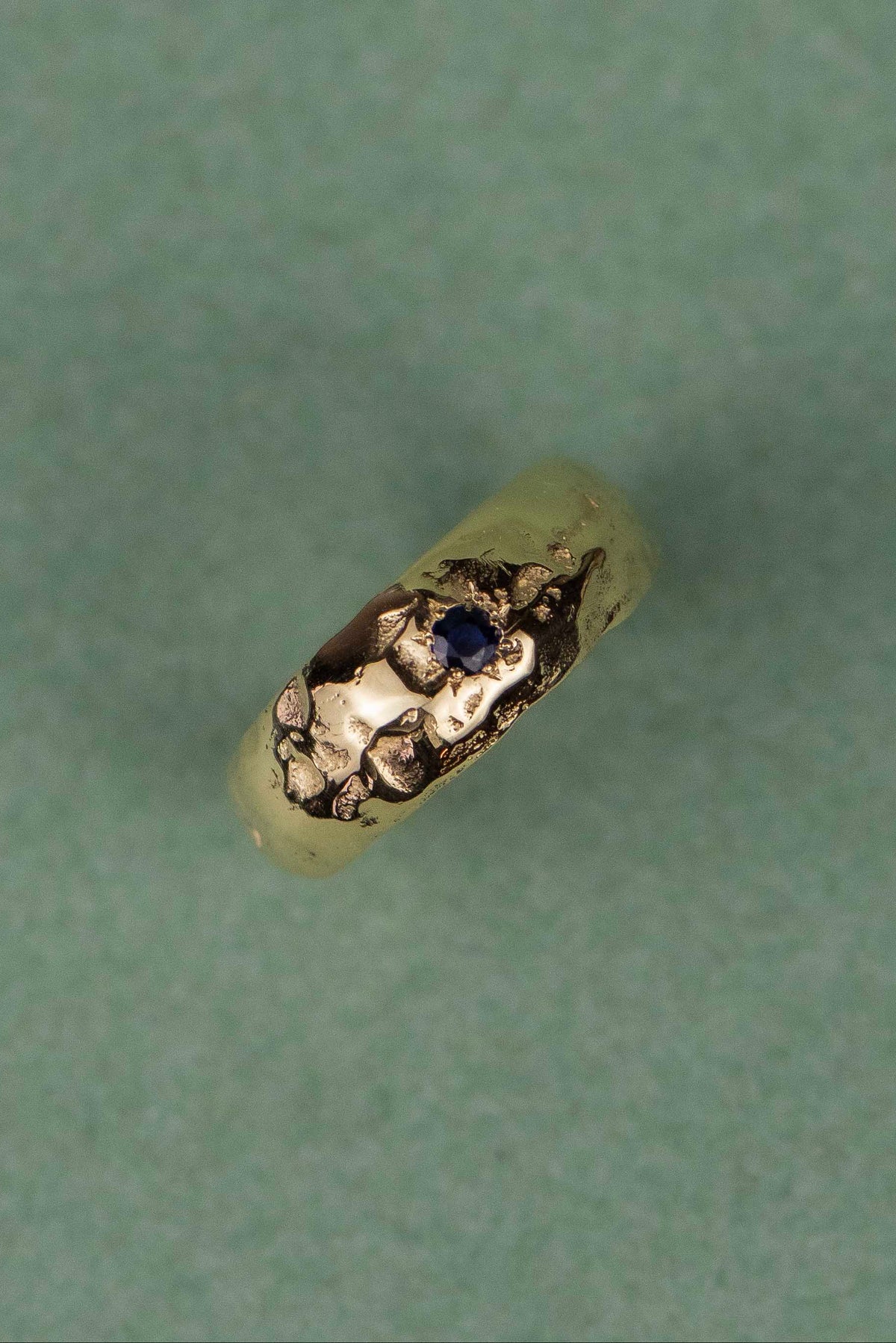 A small gold ring with a textured, hammered finish and a tiny dark blue stone, shown on a textured sage green background.